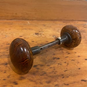 May include: A vintage door handle featuring two rounded, dark brown wooden knobs connected by a metal rod. The knobs display a distinctive wood grain pattern. Suitable for a cabinet or door.