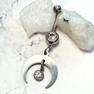 May include: A silver-toned curved barbell belly ring featuring a top ball, a middle bezel-set clear round gemstone, and a dangling crescent moon charm with a small clear round gemstone suspended from the center of the moon.