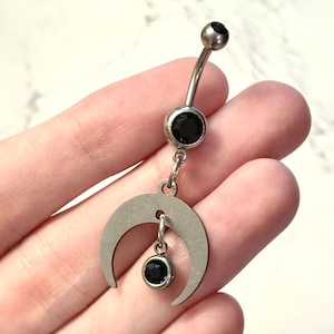 May include: A silver-toned curved barbell belly ring featuring two round black gemstones. The lower portion of the jewelry includes a dangling crescent moon charm with a small, round black gemstone suspended from the center of the moon.