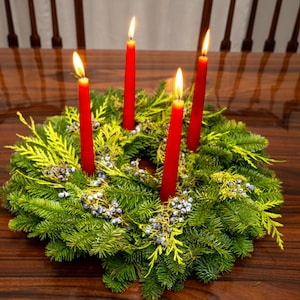 Pode incluir: Uma coroa do Advento com quatro velas vermelhas acesas, cada uma com uma chama brilhante. A coroa é feita de folhagem verde e amarela fresca, com pequenas bagas azuis, sobre uma mesa de madeira. Uma decoração de Natal.