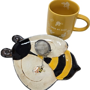May include: A yellow ceramic mug with the text "Need My Buzz" and a bee design. The mug is paired with a ceramic bee-shaped tea infuser and a matching saucer.