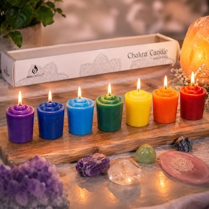 May include: A set of seven lit, unscented Chakra Candles in a row, each a different color: purple, blue, light blue, green, yellow, orange, and red. The candles are on a wooden board, with a box labeled "Chakra Candle" behind them. Crystals and a decorative plate are in front.