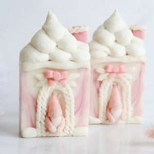 May include: Two pink and white decorative soap bars shaped like houses with white frosting-like roofs and pink ribbon accents.