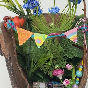 May include: A miniature fairy garden scene in a broken terracotta pot with green plants, blue flowers, a pink flower, and a colorful fabric banner.