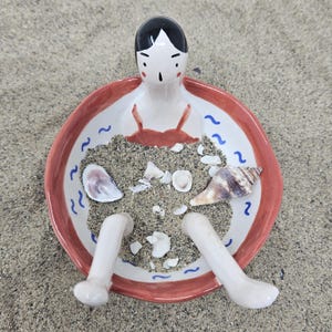 May include: A ceramic decorative bowl featuring a figure with black hair and red accents, filled with sand and seashells. The bowl has a red rim and blue wave designs. The figure's legs extend from the bowl.