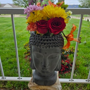 May include: A gray stone Buddha statue with a serene expression, holding a bouquet of colorful flowers in a pot. The flowers include yellow, red, pink, and orange blooms.