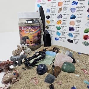 May include: A Spirit Animal Gem Hunt kit with a jar, a black scoop, and various gemstones and animal figurines on sand. The jar has a label with an elephant and sunset design. A chart with gemstone names is in the background.