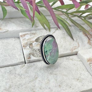 May include: A silver adjustable ring with an oval-shaped ruby zoisite stone. The stone features green and pink hues, and is framed by a twisted rope design. The ring band has a textured pattern.