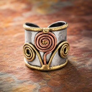 May include: A wide band ring featuring a hammered silver-tone finish with a central copper-colored wire spiral and two smaller gold-tone wire spirals on either side. The top and bottom edges are trimmed with a textured gold-tone metal border.