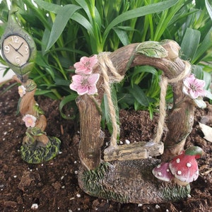 May include: A miniature fairy garden set with a clock tower, a swing set, and a mushroom. The clock tower is made of wood and has a green leaf design. The swing set is made of wood and has a rope swing. The mushroom is red with white spots.