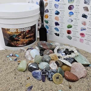 May include: A collection of gemstones in various colors and shapes, including amethyst, obsidian, and quartz, displayed on a sandy surface. A white bucket with "GEM DIGGERS" text and a black scoop are also present, along with a chart of gemstones.