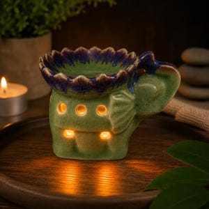 May include: Green ceramic elephant oil warmer with a blue and green floral design on top. The warmer has several holes for light to shine through. A lit candle is visible in the background.