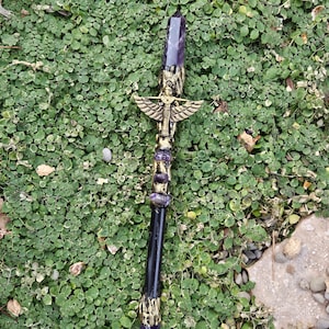 May include: A decorative staff with a large, purple crystal point. The staff features gold-colored metal accents, including a winged figure and decorative bands with purple stones. The handle is black with gold detailing. The staff is set against a backdrop of green foliage.