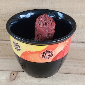 May include: A black ceramic bowl with a colorful design featuring the symbol for the sacral chakra. The bowl contains a red and brown smudge stick.