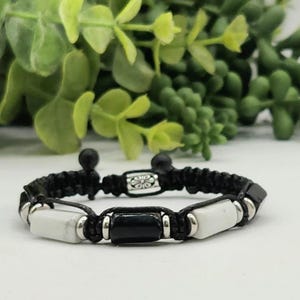 May include: A black braided bracelet with white and black rectangular beads. The bracelet has a sliding knot closure and is displayed on a white surface with green foliage in the background. The bracelet is a piece of jewelry.