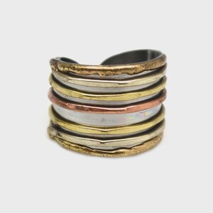 May include: A multi-layered ring with a stacked design. The ring features bands of varying colors, including gold, silver, and copper. The ring has a textured, uneven surface, giving it a rustic, handcrafted appearance. The ring is a statement piece of jewelry.