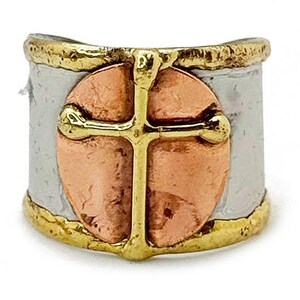 May include: A wide ring with a gold cross design on a copper-colored oval, set against a silver band. The edges are trimmed with gold, creating a layered effect. The ring has a textured appearance.