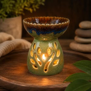 May include: A green ceramic essential oil diffuser with leaf-shaped cutouts. The top rim is brown and blue. A lit candle is inside, casting a warm glow. The diffuser sits on a wooden tray, suggesting a spa or relaxation theme.