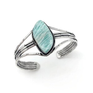May include: A silver cuff bracelet featuring a large, oval-shaped, light blue gemstone. The stone is set in a black-lined bezel and is the focal point of the bracelet. The bracelet has multiple parallel bands and a hammered texture.
