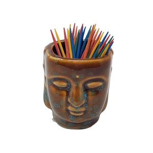 May include: Brown ceramic toothpick holder shaped like a Buddha face with a blue glaze. The holder is filled with colorful wooden toothpicks.