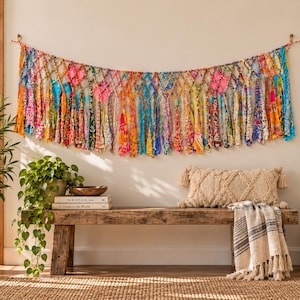 May include: A wall hanging made of multicolored fabric strips tied in a knotted net pattern. The textile garland features a vibrant array of floral and geometric prints in shades of pink, orange, blue, yellow, and green, suspended by a cord.