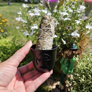 May include: A white sage smudge stick in a black ceramic holder with a pentagram design.