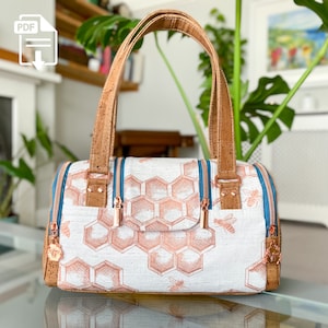 Könnte beinhalten: Eine weiße und braune Tragetasche mit einem Wabenmuster und Bienen. Die Tasche hat Korkgriffe und roségoldene Reißverschluss-Zipper.