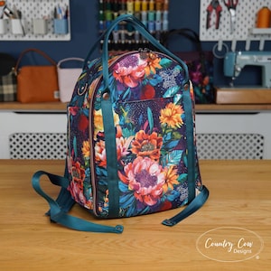 May include: A floral print backpack with teal straps and handles. The bag features a vibrant pattern of pink, orange, and blue flowers against a dark background. The bag has a zippered closure and a front pocket. The brand name "Country Cow Designs" is visible.
