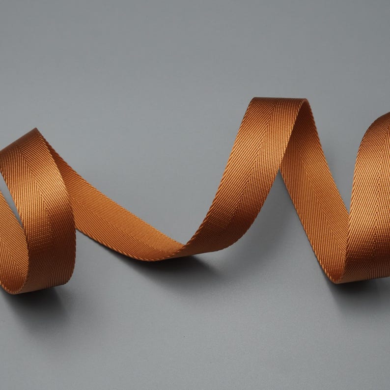 Peut inclure: Gros plan d'un ruban de couleur cuivre avec un motif de tissage en chevrons. Le ruban est enroul&eacute; et repose sur un fond gris. La texture et la couleur du ruban sont mises en &eacute;vidence dans l'image.