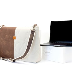 Op de afbeelding: Een bruine en witte schoudertas met een bijpassende riem en een laptop. De tas heeft een bruin paneel aan de linkerkant en een wit gewatteerd paneel aan de rechterkant. De laptop is zwart.