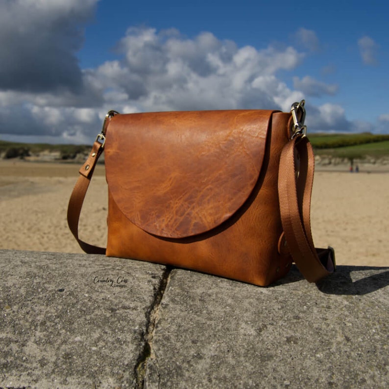 May include: A brown leather crossbody bag with a flap closure and a long adjustable strap. The bag is sitting on a rock with a sandy beach in the background.