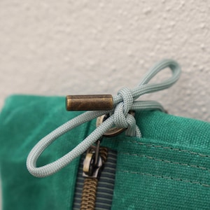 Peut inclure: Gros plan sur un sac en toile vert sarcelle avec une fermeture éclair et une tirette en laiton. La tirette est une corde bleu clair nouée en boucle. L'extrémité de la tirette est en laiton décoratif.