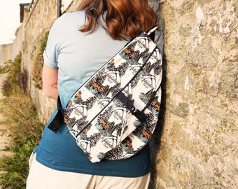 May include: A white and black sling bag with a repeating pattern of a jeep, trees, and the word "Adventure". The bag has a black strap and a small black pocket. The bag is worn over the shoulder.