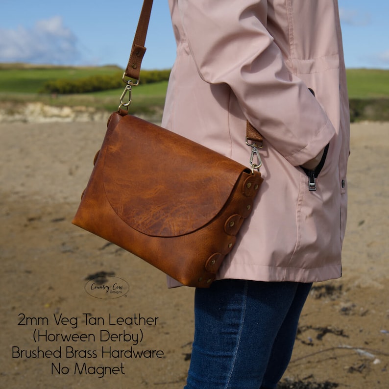 May include: A brown leather crossbody bag with a flap closure and brushed brass hardware. The bag is made of 2mm thick Horween Derby leather and has no magnet closure. The text "Country Cow Designs" is visible on the bag.