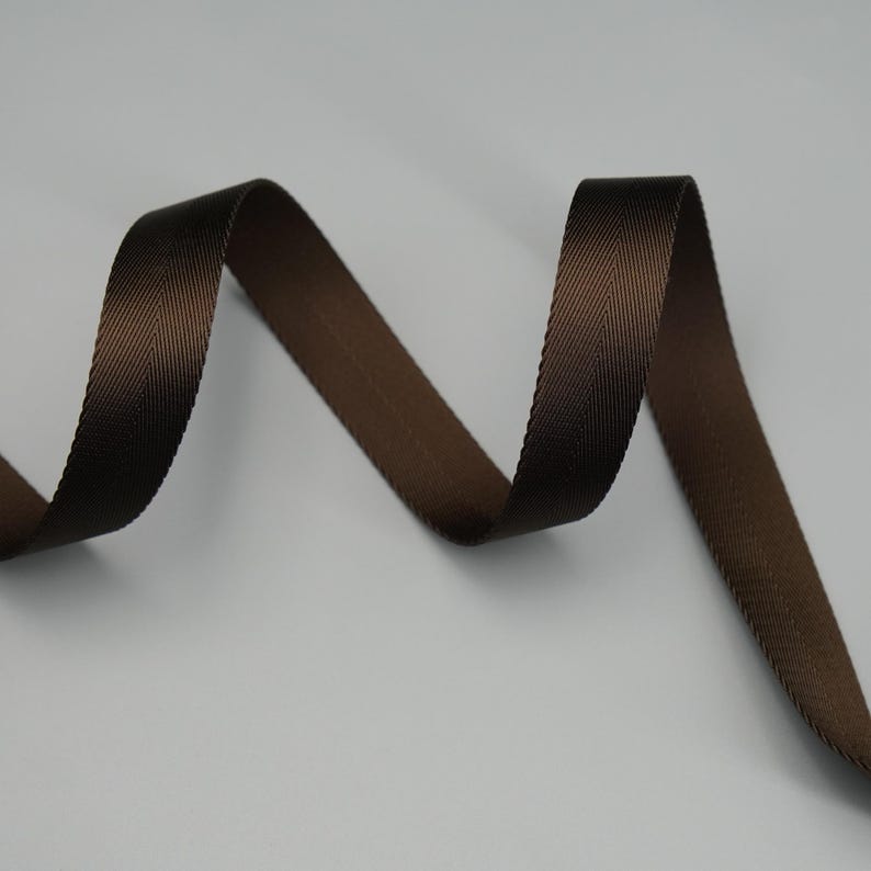 Peut inclure: Gros plan sur un ruban marron avec un motif chevron subtil. Le ruban est d'un brun riche et fonc&eacute; et semble &ecirc;tre fait d'un mat&eacute;riau durable. Le ruban est incurv&eacute; et a un l&eacute;ger &eacute;clat, sugg&eacute;rant qu'il pourrait &ecirc;tre utilis&eacute; pour divers travaux manuels ou d&eacute;coratifs.