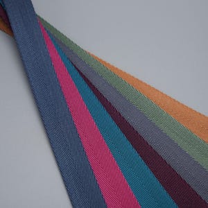 Peut inclure: Une collection de bandes de tissu à motif chevrons colorées dans les tons de bleu, rose, sarcelle, violet, vert et orange. Les bandes sont disposées en diagonale sur un fond gris.