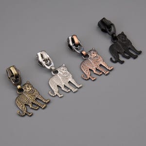 Puede incluir: Cuatro tiradores de cremallera metálicos con diseños de leopardo en acabados bronce, plata, cobre y negro. Cada tirador presenta una silueta detallada de leopardo unida a un cursor de cremallera.