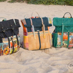 May include: Three colorful backpacks with travel-themed designs are displayed on a sandy beach. The backpacks feature various colors, including black, cork, and green, with travel destination imagery on the pockets and straps. Each bag has adjustable straps and a top handle.