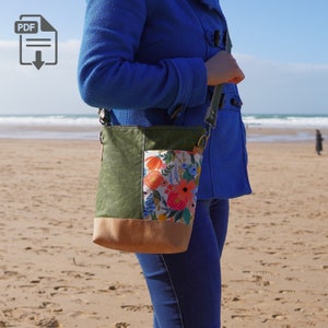 May include: A green, brown, and floral patterned crossbody bag with a long strap. The bag is worn by a person on a beach.