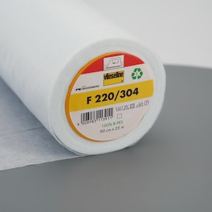 Puede incluir: Un rollo blanco de tejido de entretela Vlieseline F 220/304. La etiqueta es amarilla con texto negro y un código de barras. La etiqueta también incluye instrucciones de cuidado y el texto "100% R-PES 90 cm x 25 m".