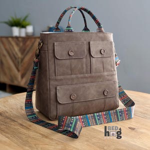 Peut inclure: Un sac fourre-tout en cuir marron avec plusieurs poches et une bandoulière à motif coloré. La bandoulière présente un motif de livres empilés.