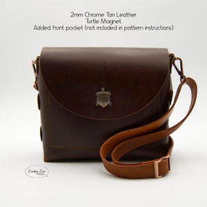 May include: A brown leather crossbody bag with a flap closure and a turtle magnet. The bag has a brown leather strap and a front pocket. The bag is made of 2mm chrome tan leather.