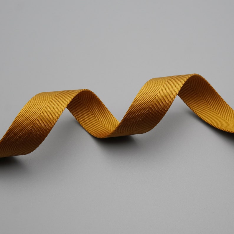 Peut inclure: Un ruban jaune dor&eacute; en tissage chevrons, ondulant sur un fond gris. La texture du ruban est visible, avec un l&eacute;ger &eacute;clat. Ce ruban convient &agrave; divers projets d'artisanat et de couture.