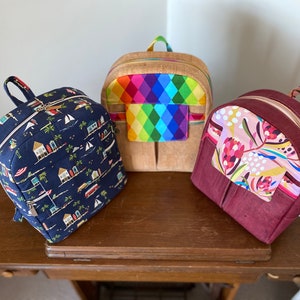 May include: Three different backpacks with unique fabric patterns. The first backpack is navy blue with a white and yellow beach scene. The second backpack is brown with a rainbow diamond pattern. The third backpack is burgundy with a pink and white floral pattern.