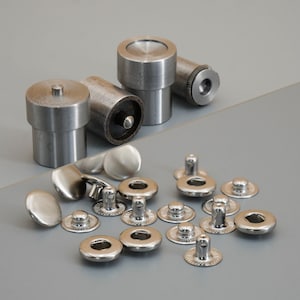 May include: A collection of metal tools and fasteners. The tools are cylindrical with flat tops and bottoms. The fasteners include round, flat pieces and pieces with a central post and a decorative edge. The items are silver and sit on a gray surface.