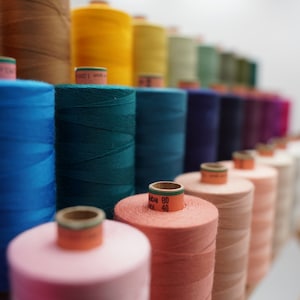 May include: A close-up of a variety of spools of thread in different colours, including blue, green, pink, and orange. The spools are arranged in a row, with the closest spool being a light pink colour.