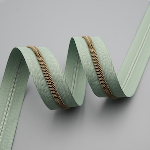 May include: A close-up of a sage green zipper with a brass zipper pull. The zipper is coiled in a spiral shape.