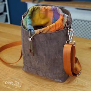 Puede incluir: Un bolso tipo cubo marrón con cierre de cordón y forro de tela a cuadros multicolor. El bolso incluye una correa de hombro ajustable de color naranja brillante sujeta con herrajes de metal cobrizo y un cierre de cordón.