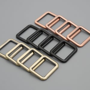 May include: Three sets of square metal rings in gold, black and rose gold. The rings are stacked on top of each other.