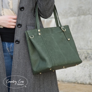 Pode incluir: Uma bolsa tote verde com ferragens douradas, incluindo rebites na parte inferior e detalhes nas alças. A bolsa tem um design estruturado e é carregada por uma pessoa que usa um casaco cinza. O texto "Country Cow Designs" é visível.